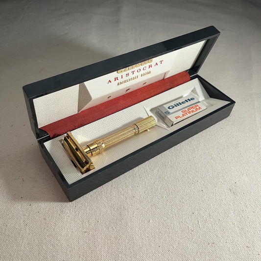 Maquinilla de afeitar Gillette Aristocrat dorada de 1962 en su estuche negro abierto, con tarjeta y paquete de cuchillas.