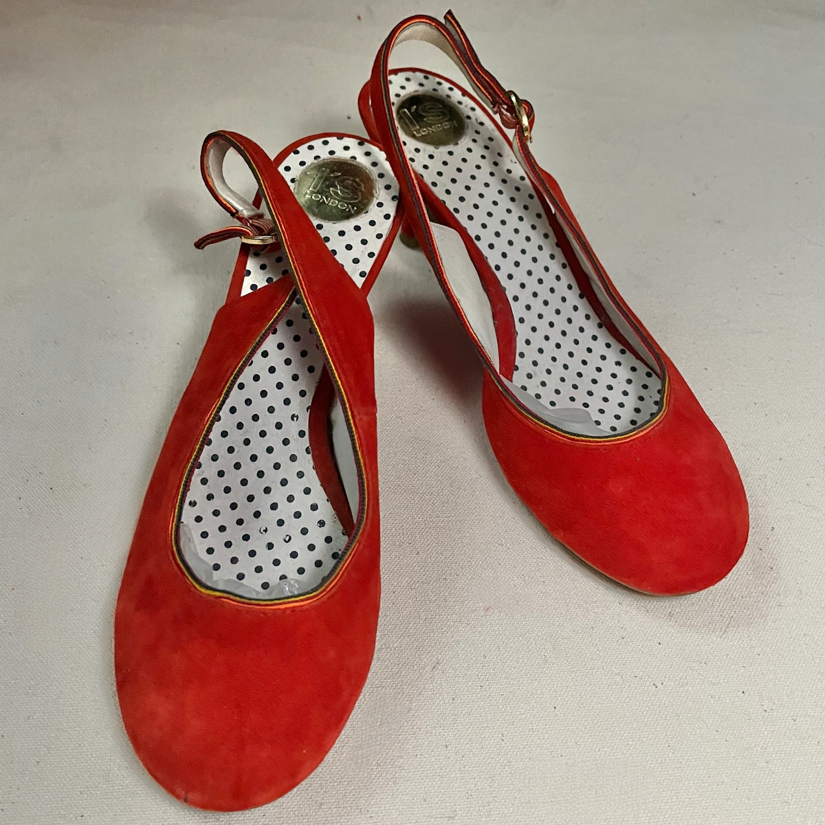 Par de zapatos destalonados en ante rojo con tacón de carrete, tira de talón con hebilla dorada y plantilla blanca de lunares negros, vistos de tres cuartos.