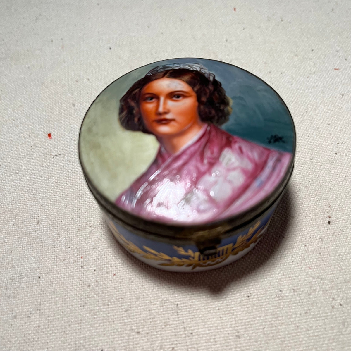 Cajita redonda de porcelana Bidasoa con retrato de maja pintado a mano en la tapa, cerco dorado y banda decorativa con laurel y ánfora sobre fondo azul.