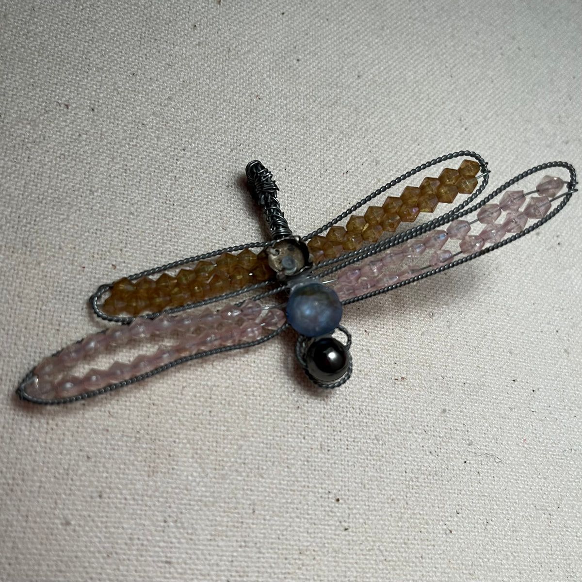 Broche de libélula en alambre plateado con alas de abalorios de vidrio en topacio dorado y rosa pálido, y cuerpo con perlas de cristal gris y hematite, sobre lino claro.