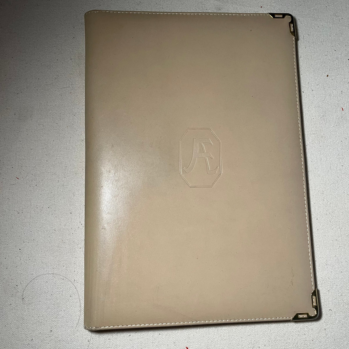 Carpeta-agenda de piel beige con esquineras metálicas doradas y monograma AE grabado en la cubierta, abierta mostrando el interior con bolsillos y sujetador de bolígrafo.