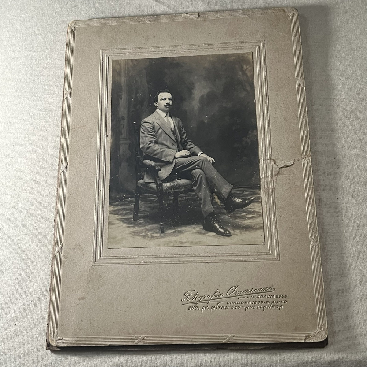 Retrato de estudio de un hombre con bigote, sentado y con las piernas cruzadas, montado en cartón con paspartú en relieve y el sello de Fotografía Americana, Buenos Aires